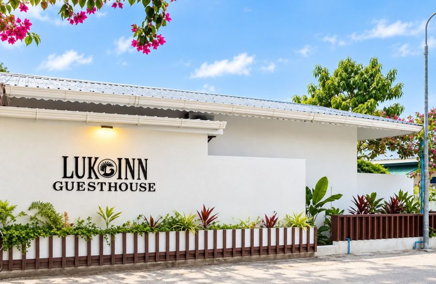 White single-storey building with the Luk Inn Guesthouse sign, a planted fence and pink bougainvillea