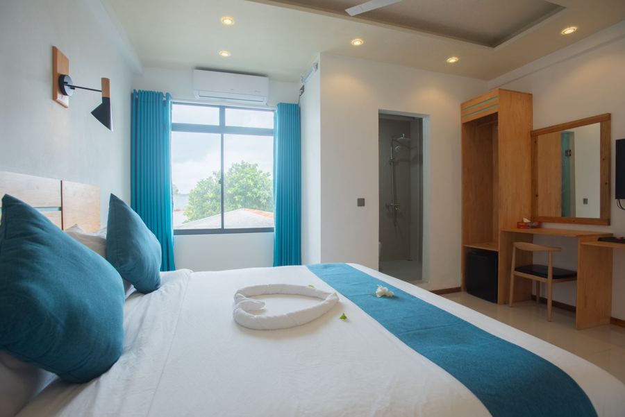 Bed with teal cushions and runner by a large window with blue curtains, desk and mirror behind