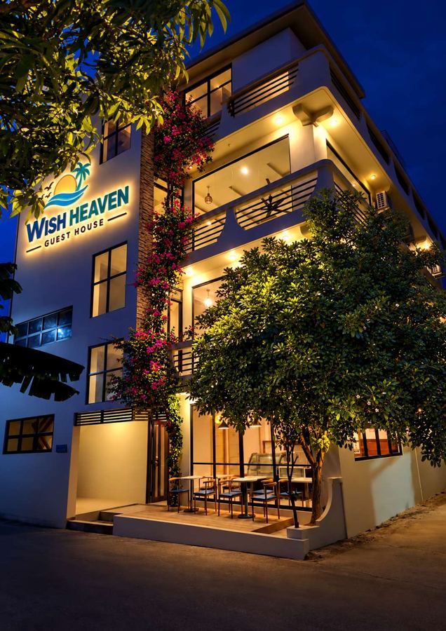 Four-storey Wish Heaven Guesthouse lit up at night, with its sign, flowering bougainvillea and a café terrace at street level